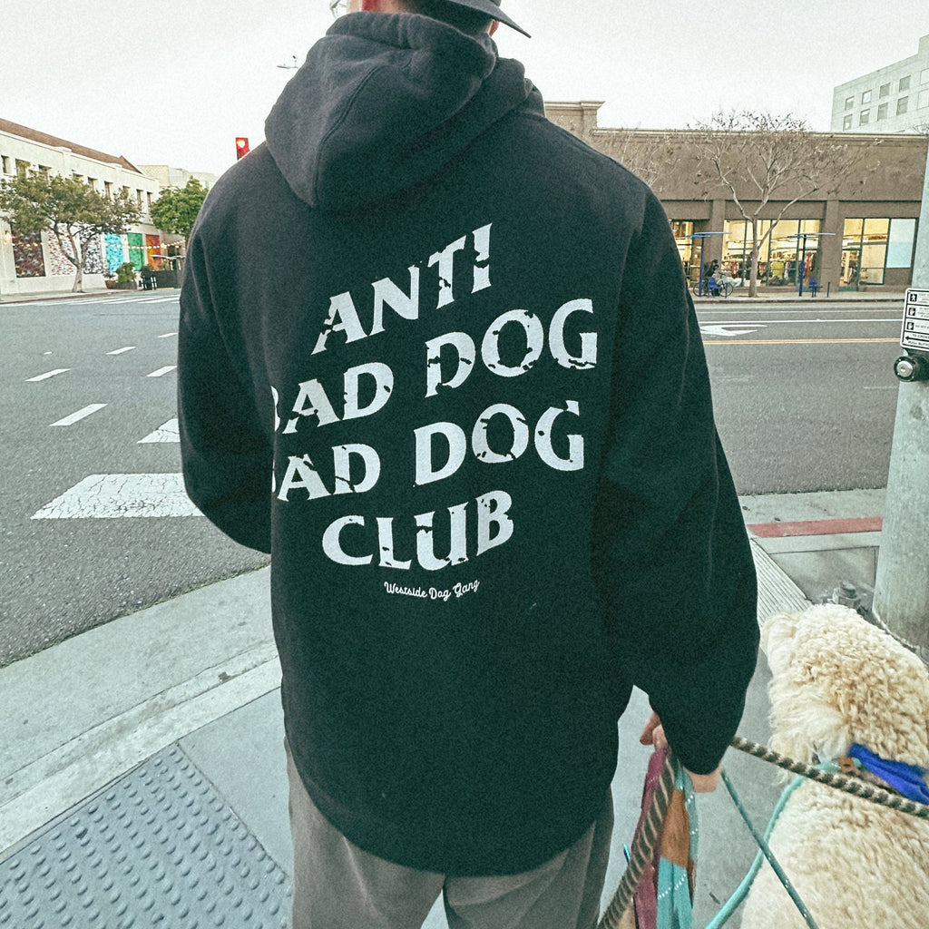 A confident man walks his dogs down a bustling downtown street, proudly wearing an 'Anti Bad Dog Bad Dog Club' hoodie.