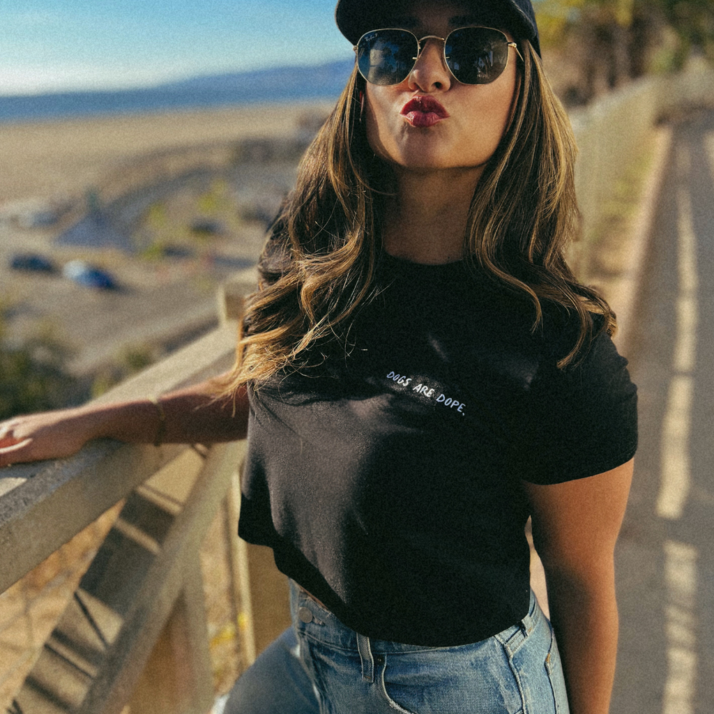 A model stands on a cliff, gazing at the beach with a playful kissing face, wearing a Dogs Are Dope shirt and hat, exuding coastal chic vibes.