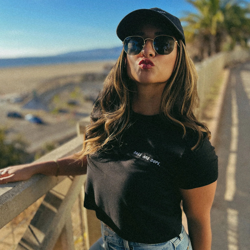 A model stands in a park overlooking the ocean in Santa Monica, wearing a Dogs Are Dope shirt and hat, while making a playful kissing face.